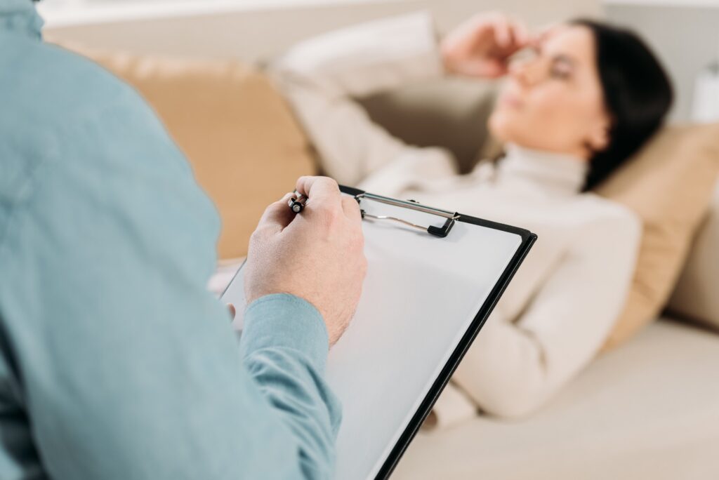 cropped shot of psychotherapist writing on clipboard and young woman lying on couch in office